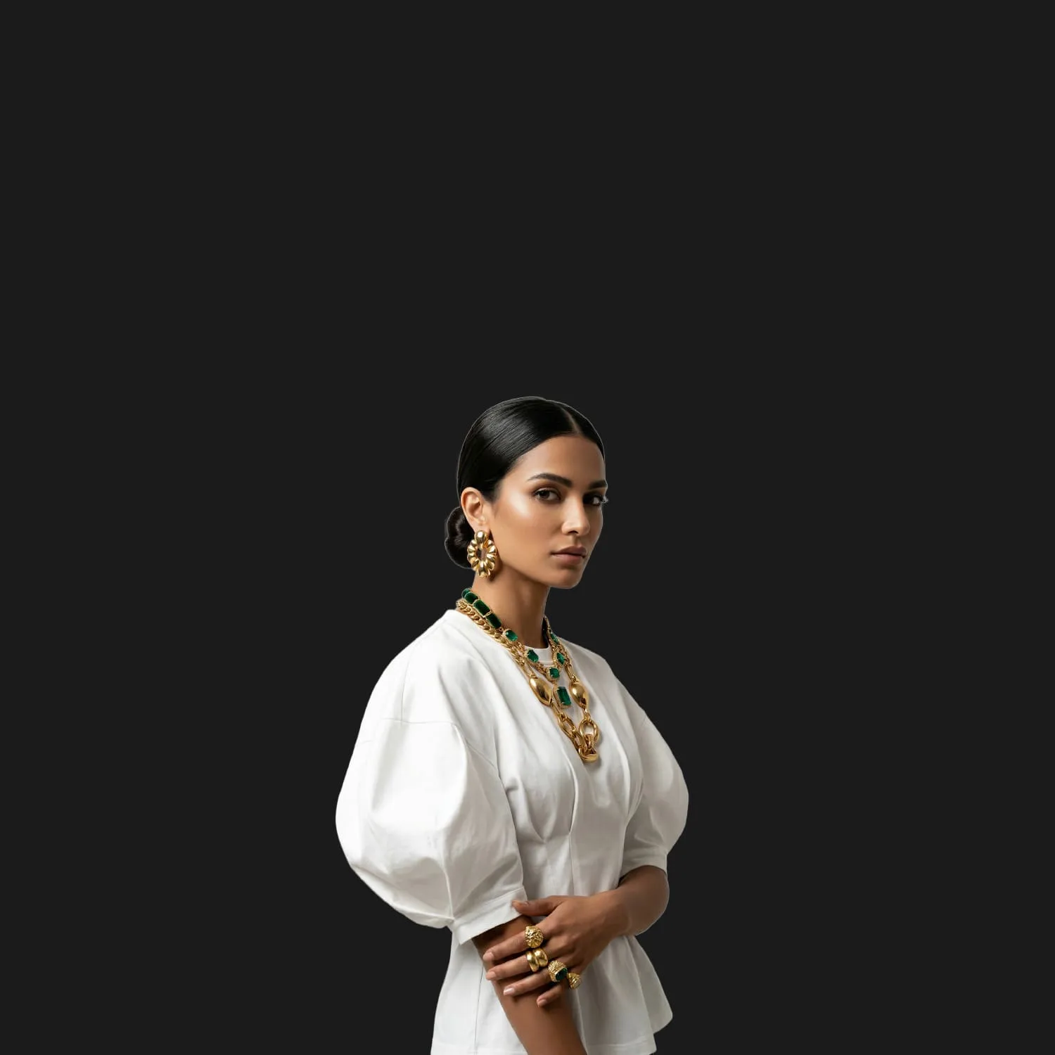 Model in a white puff sleeve top layered with gold chains and emerald necklaces on black