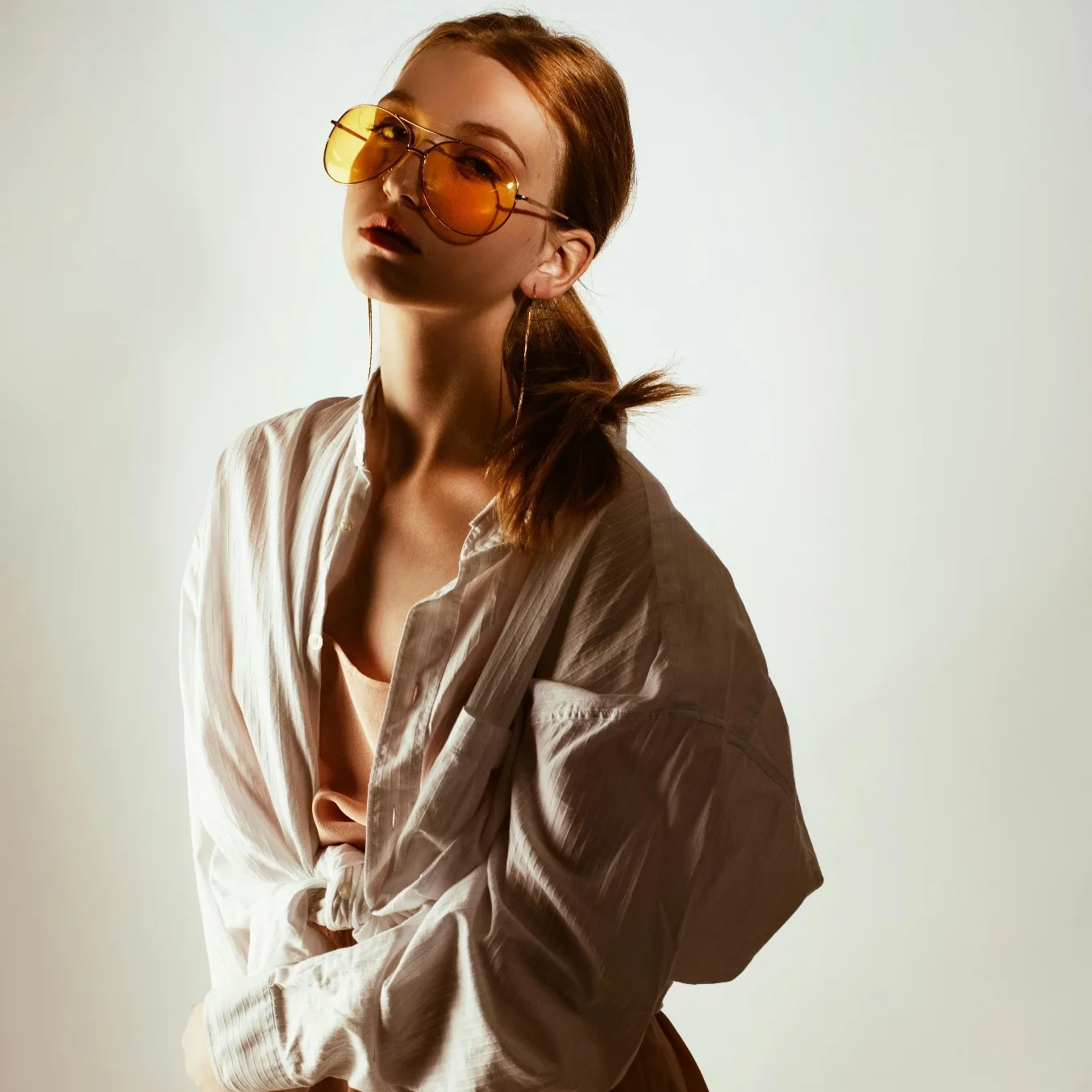 Model in yellow tinted aviator sunglasses and an oversized white shirt posing in a studio