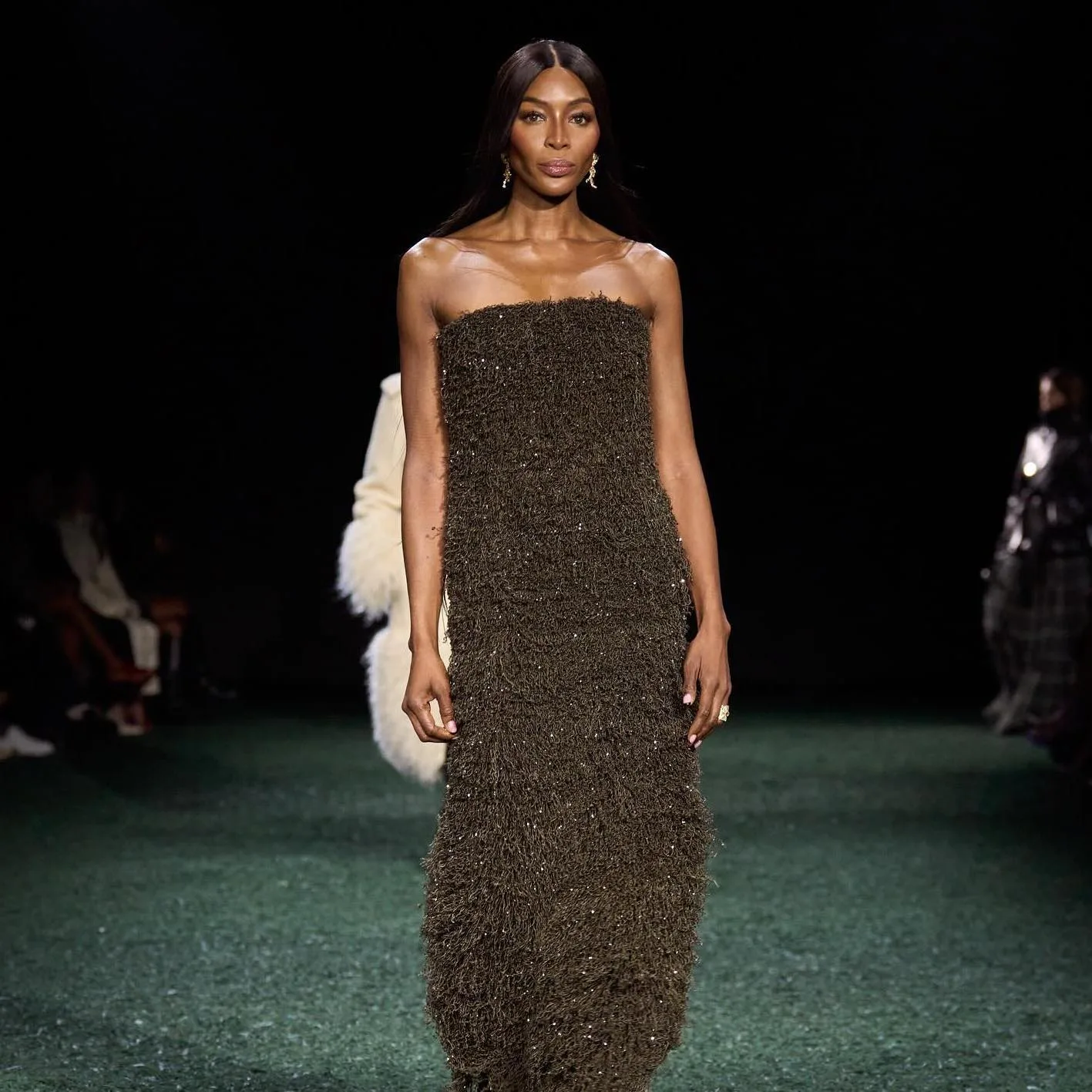 A model walking a London Fashion Week runway in a long strapless dress with a dense fringed texture