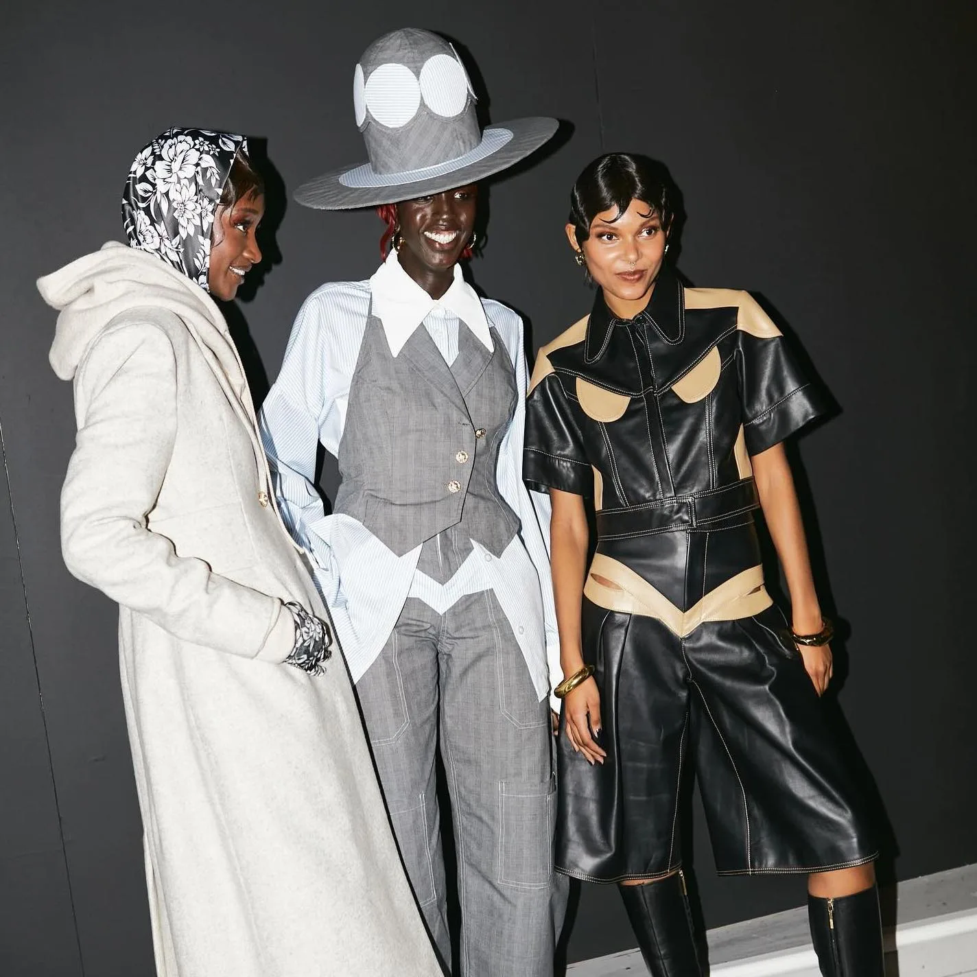 Three models backstage at London Fashion Week in a cream coat, a gray tailored look with a wide brimmed hat, and a black leather outfit