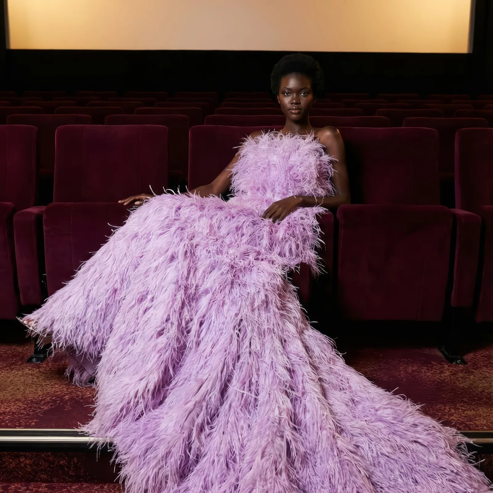 Model in a lilac feather gown seated across red velvet seats in an empty cinema