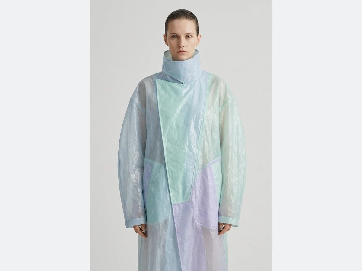 A model in an iridescent coat, photographed against a plain light grey studio wall